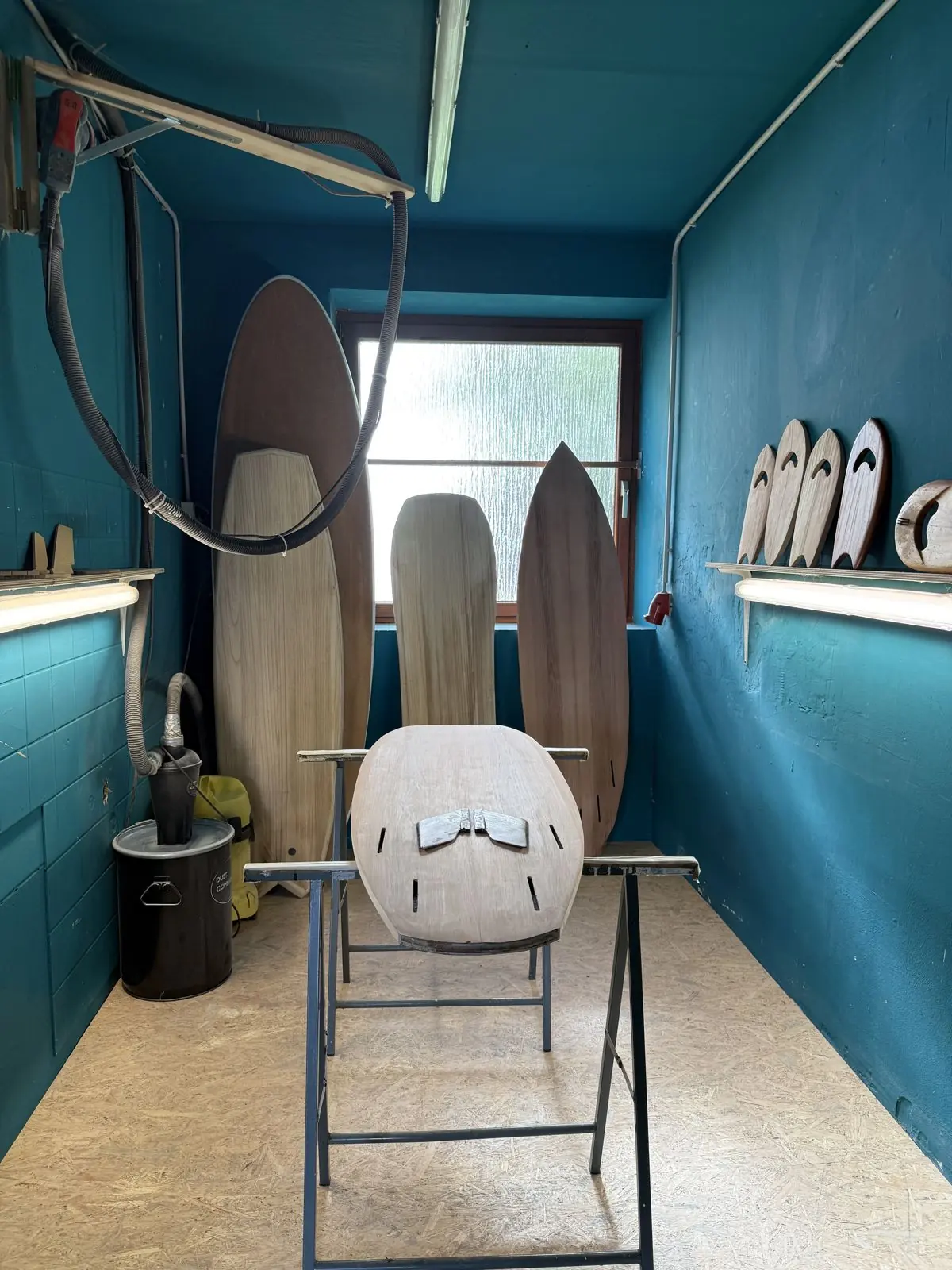 Holzsurfbrett mit Finnenkästen auf Shaping-Böcken, dahinter drei Holzbretter vor dem Fenster, Handplanes auf dem Wandbord