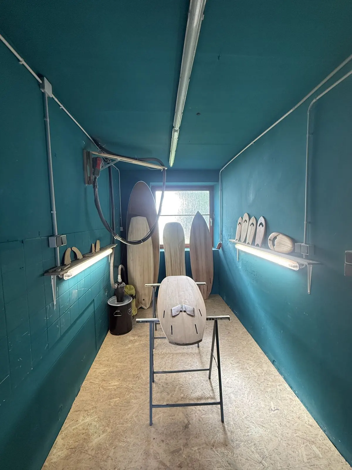 Schmaler petrolfarbener Shape-Raum mit Seitenlicht, Absaugung von der Decke und einem Holzsurfbrett auf Shaping-Böcken
