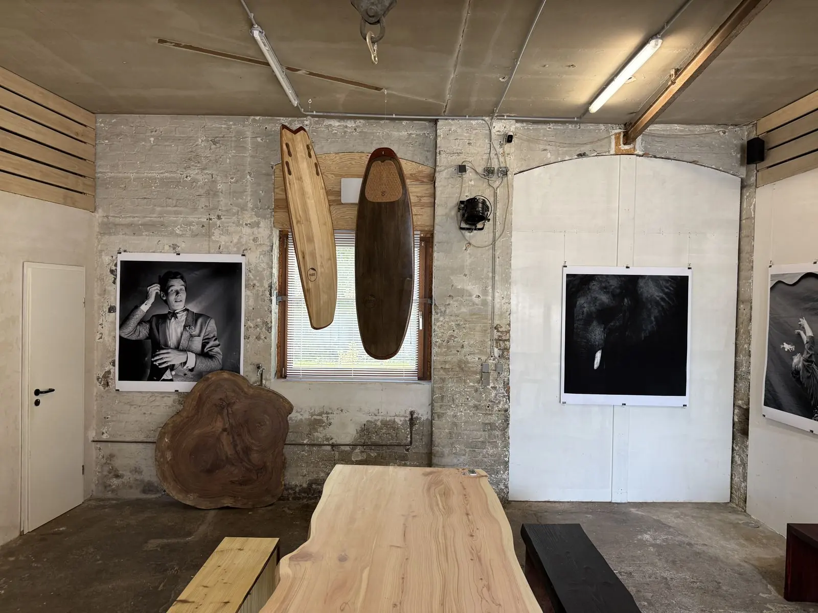 Ausstellungsraum mit zwei hängenden Holzsurfbrettern, Baumscheibe, Holztisch und Schwarzweiß-Fotografien